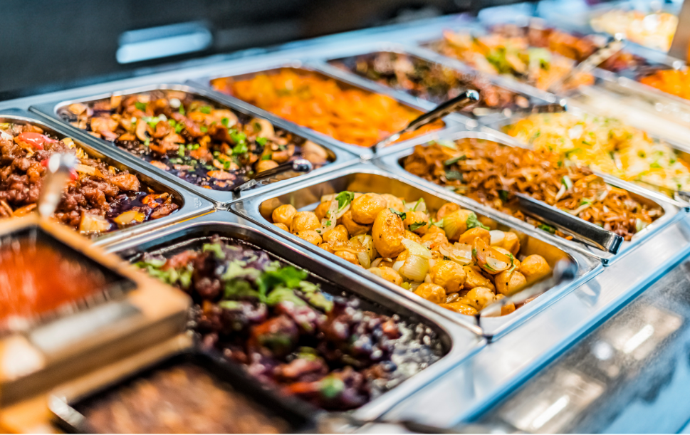 Hot food bar with multiple trays of prepared dishes, allowing customers to view and choose meal options.